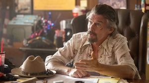 A cut up Ethan Hawke in a western-style shirt sits behind an office desk. A western had with sunglasses on the brim sits on the desk in front of him.