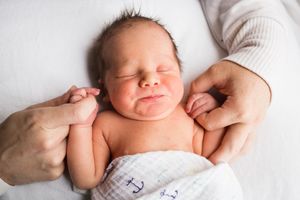 Baby Owen at two days old, eyes closed, mouth in a subtle grin, holding his mom and dad's fingers